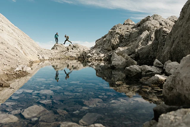 Two hikers on rocky terrain near the Valluga in the Austrian Alps, one leaping between rocks, reflected in a clear alpine mountain pool in the foreground.